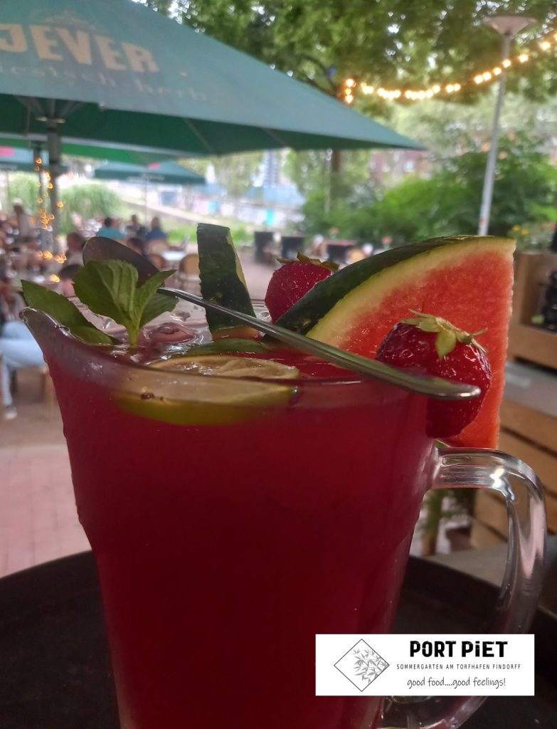 Ein großes Glast mit einer roten Flüssigkeit, einer Melonenschnitte, Früchten und einem Strohhalm unter einem Sonnenschirm. In einer Ecke das Logo des Sommergartenlokals Port Piet.